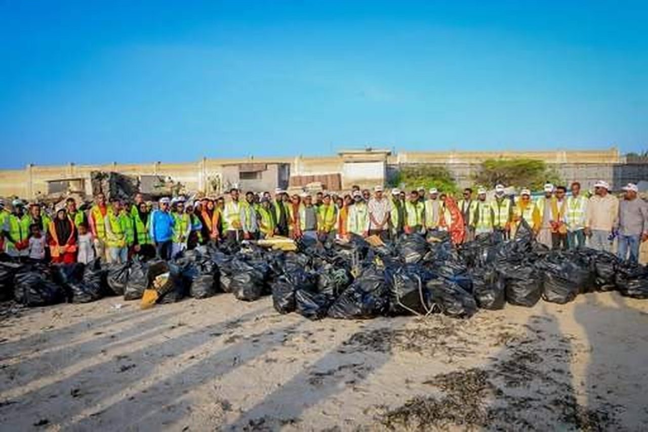 A long line of volunteers in safety vests behind rows of black bags of rubbish on a beach.